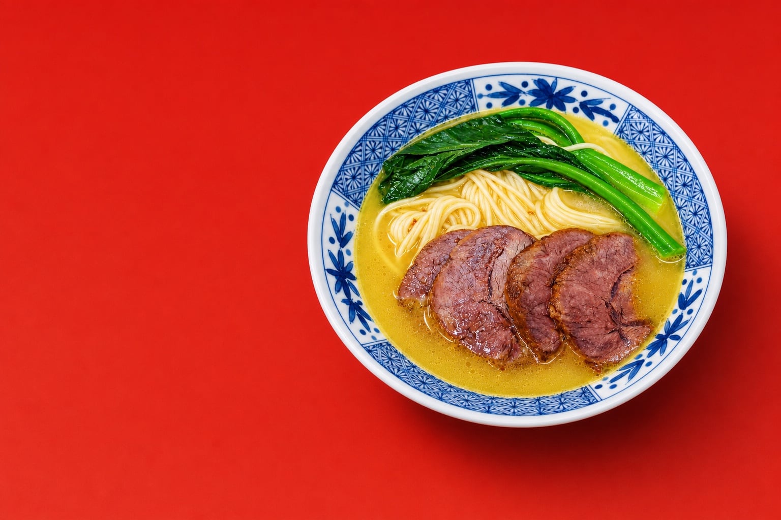 A blue-and-white bowl of Consort Yang’s Chicken Noodle — sliced braised beef, wheat noodles and choy sum in golden broth — on a red field